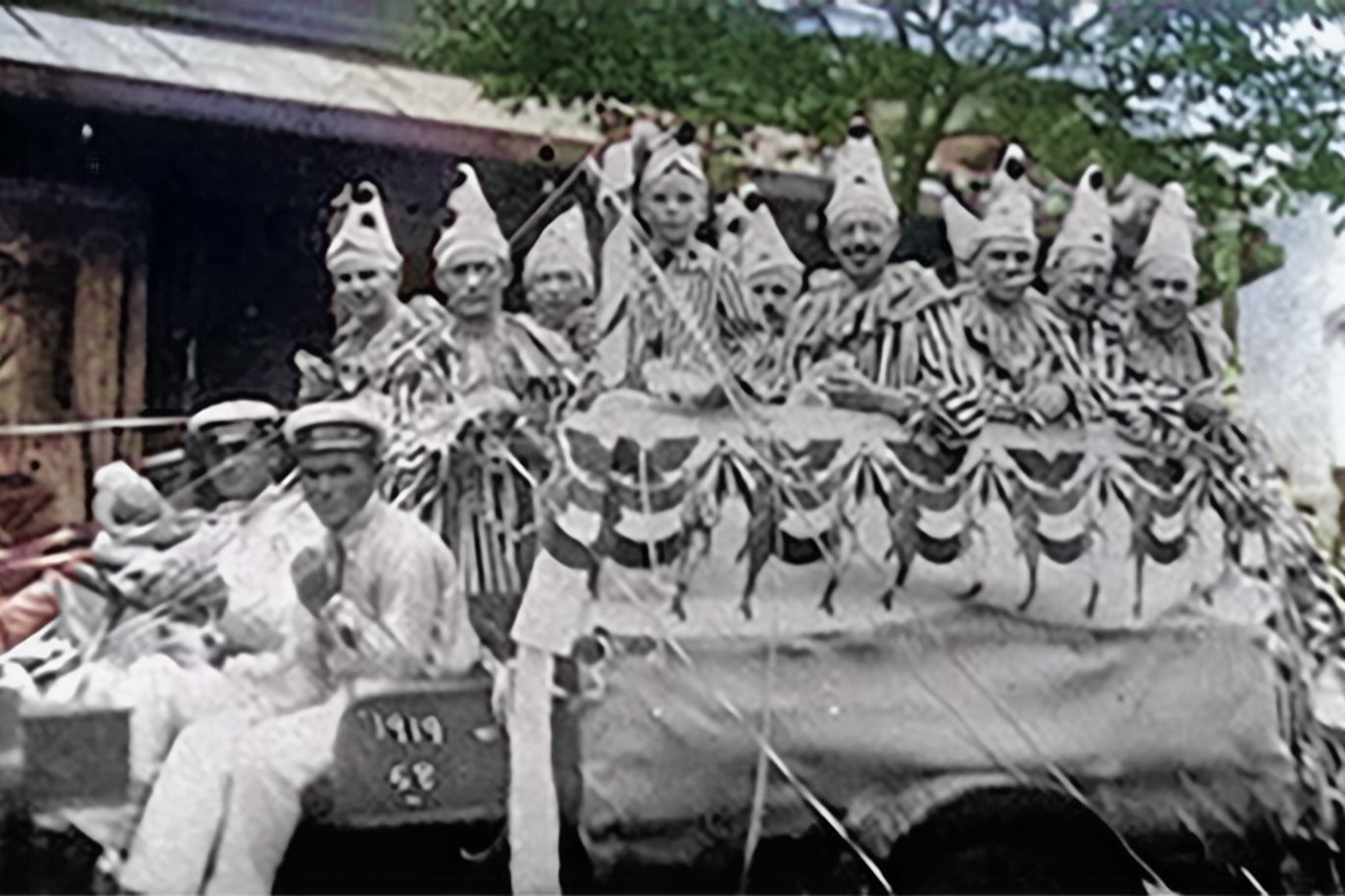 Corso carnavalesco with cars populated by revelers in costumes, decorated with streamers. Colorized reproduction.